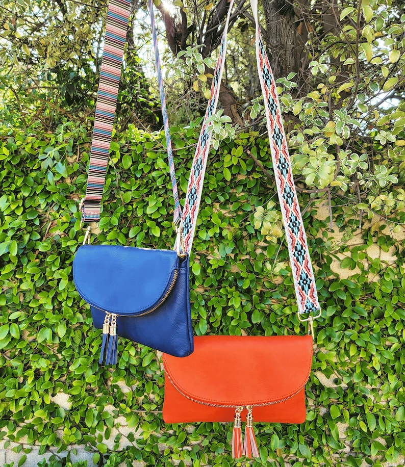 Pu&ograve; includere: Due borse a tracolla, una blu reale e una arancione, ciascuna con patta, cerniera dorata e due nappe abbinate. La borsa blu ha una tracolla a righe multicolore, mentre la borsa arancione ha una tracolla bianca con un motivo geometrico.