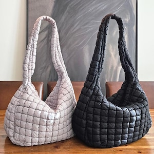 May include: Two quilted tote bags. One is a light gray color, and the other is black. Both bags have a quilted pattern and a long, padded shoulder strap. The bags are sitting on a wooden table.