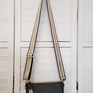 Pu&ograve; includere: Una borsa a tracolla rettangolare nera con texture intrecciata e chiusura superiore a zip. La borsa presenta una lunga tracolla regolabile con un motivo intrecciato color crema e oro, bordi neri e dettagli in metallo color oro.