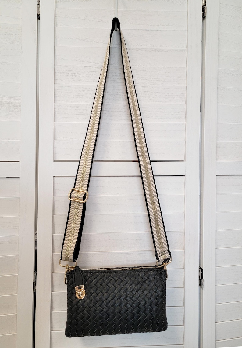 Pu&ograve; includere: Una borsa a tracolla rettangolare nera con texture intrecciata e chiusura superiore a zip. La borsa presenta una lunga tracolla regolabile con un motivo intrecciato color crema e oro, bordi neri e dettagli in metallo color oro.