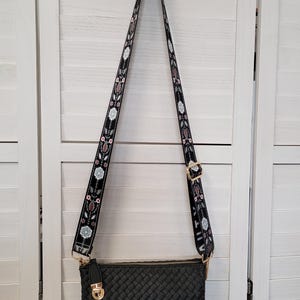 Pu&ograve; includere: Una borsa a tracolla rettangolare nera con trama intrecciata e chiusura in metallo dorato. La borsa presenta una lunga tracolla regolabile decorata con un motivo floreale ripetuto in bianco, rosa e marrone su sfondo nero.