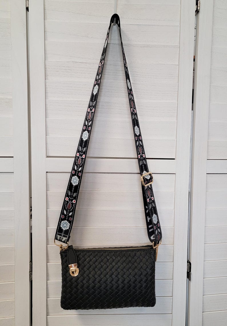 Pu&ograve; includere: Una borsa a tracolla rettangolare nera con trama intrecciata e chiusura in metallo dorato. La borsa presenta una lunga tracolla regolabile decorata con un motivo floreale ripetuto in bianco, rosa e marrone su sfondo nero.
