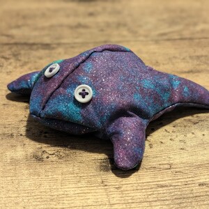 May include: A plush toy shaped like a manta ray with a galaxy print fabric. The toy has two white buttons for eyes.