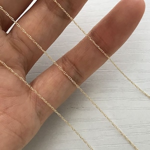 May include: Close-up of a delicate gold-coloured chain necklace. The chain is thin and appears to be made of small, interconnected links. The necklace is resting on a light-coloured surface.