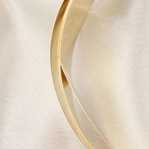 May include: A close-up shot of a gold-coloured, curved object with a layered design. The object has a smooth, reflective surface and appears to be made of metal. The background is a soft, off-white fabric, creating a contrast with the gold.
