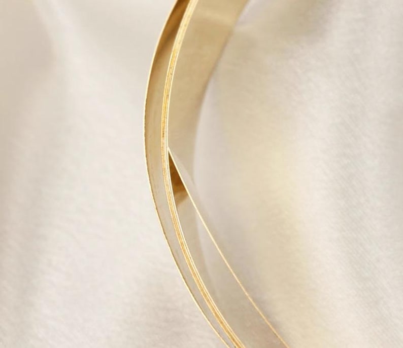 May include: A close-up shot of a gold-coloured, curved object with a layered design. The object has a smooth, reflective surface and appears to be made of metal. The background is a soft, off-white fabric, creating a contrast with the gold.