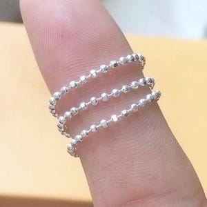 May include: A silver ring featuring three bands of tiny, linked beads. The ring is worn on a finger, highlighting the intricate design and the reflective quality of the metal. The ring's understated elegance makes it a versatile piece of jewellery.