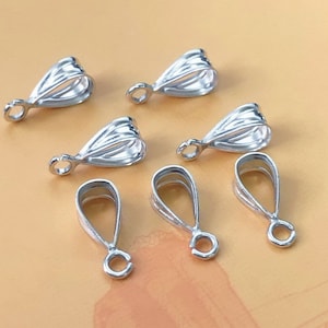 Puede incluir: Seis baúles de metal de color plateado con forma de lágrima y un bucle en la parte superior. Los baúles están dispuestos de forma dispersa sobre una superficie de color marrón claro.