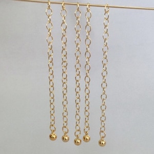 Pode incluir: Cinco correntes douradas com pequenas bolas douradas na ponta. As correntes estão penduradas em um fio fino dourado.