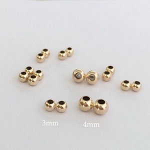 Pode incluir: Um conjunto de contas de metal douradas em dois tamanhos, 3 mm e 4 mm. As contas são redondas e têm um furo no centro. Elas estão dispostas em pares sobre uma superfície branca.