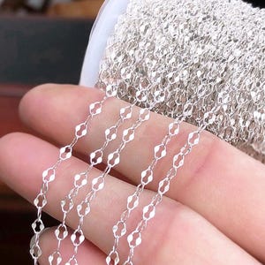 May include: Close-up of a silver-colored chain with a repeating diamond-shaped link design. The chain is partially unraveled from a larger spool, showcasing its delicate construction and reflective surface. Ideal for jewelry making.