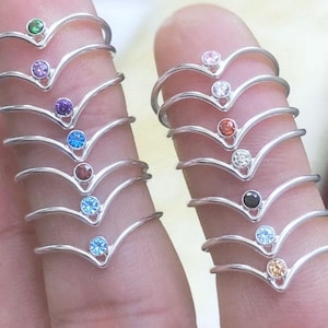 May include: A selection of silver V-shaped rings, each featuring a different coloured gemstone. Gemstones include green, purple, blue, and brown. The rings are displayed on a finger.