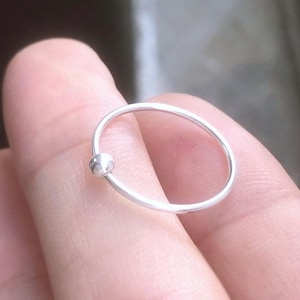Könnte beinhalten: Ein kleiner, silberner Nasenring mit schlichtem Design. Der Ring hat ein dünnes, kreisförmiges Band mit einer kleinen, runden Kugel auf einer Seite. Der Schmuck besteht aus glänzendem Metall.