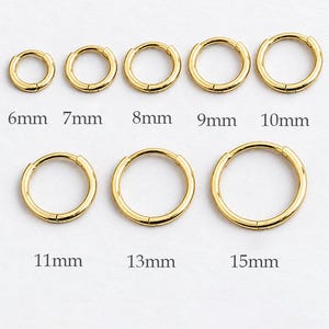 Könnte beinhalten: Eine Sammlung von acht goldfarbenen Creolen, angeordnet in zwei Reihen, mit Durchmessern von 6mm, 7mm, 8mm, 9mm, 10mm, 11mm, 13mm und 15mm. Jeder kreisförmige Ohrring hat ein schlichtes, glattes Design mit einem Klappverschluss-Mechanismus.