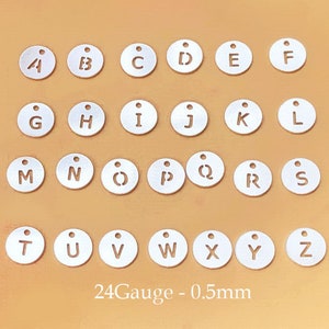 Puede incluir: Un conjunto de 26 abalorios de letras del alfabeto de metal plateado, cada uno con una letra diferente del alfabeto. Los abalorios están dispuestos en filas sobre una superficie marrón claro. El texto "24Gauge - 0.5mm" está impreso debajo de los abalorios.
