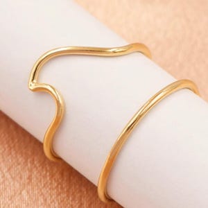 May include: Two gold rings, one with a wavy design and the other a simple band, are stacked on a white surface.