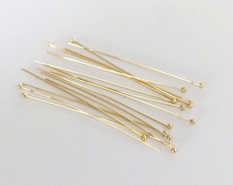 May include: A group of gold head pins with round heads. The pins are arranged in a random pattern on a white surface.