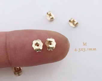 May include: Two gold-colored earring backs, measuring 4.3 x 5.1 mm, are shown on a finger. The backs are designed to secure earrings in place.