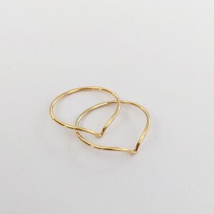 May include: Two delicate gold rings, each with a V-shaped design, are intertwined against a white background. The rings have a smooth, polished finish, reflecting light. The minimalist design suggests a focus on simplicity and elegance, suitable for everyday wear or special occasions.