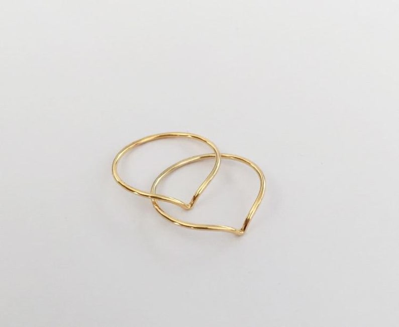 May include: Two delicate gold rings, each with a V-shaped design, are intertwined against a white background. The rings have a smooth, polished finish, reflecting light. The minimalist design suggests a focus on simplicity and elegance, suitable for everyday wear or special occasions.
