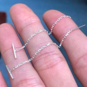 以下が含まれることがあります： 繊細なシルバーチェーンのイヤリングのペア。各イヤリングは、耳に通る細いチェーンで接続された、下部に小さな長方形のバーを備えています。イヤリングはぼやけた背景に対して表示されます。