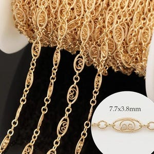 Può includere: Catena color oro con un intricato design a filigrana. La catena presenta maglie ovali con un motivo a spirale decorativo, che misurano 7,7 x 3,8 mm. La catena è mostrata in un rotolo, con diversi fili che pendono.