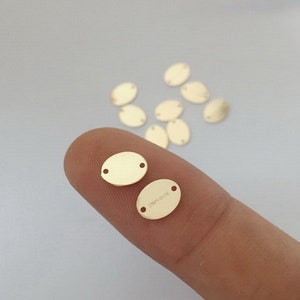 May include: A close-up of two gold oval-shaped metal blanks with two holes each. The blanks are approximately 2.5 cm long and 1.25 cm wide. The blanks are on a finger.