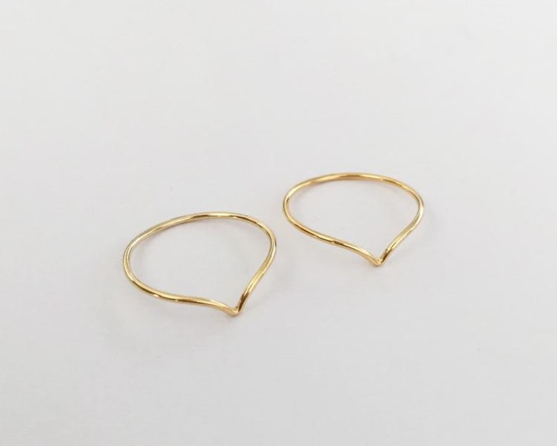 May include: Two delicate gold rings, each with a teardrop or petal shape. The rings are thin and appear to be handmade, with a slightly textured surface. The background is a plain, off-white color.