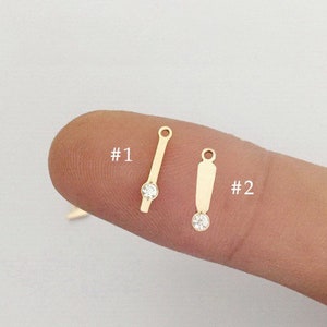 May include: Two gold-toned jewelry findings with a clear rhinestone. The finding on the left is a straight bar with a rhinestone in the center. The finding on the right is a rectangular shape with a rhinestone at the bottom. The findings are labeled #1 and #2.