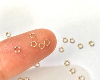 May include: A close-up of a finger holding several small gold jump rings. The rings are scattered on a white surface.