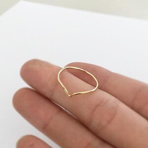May include: A delicate gold-colored ring with a V-shaped design. The ring is thin and appears to be made of a single piece of metal. The ring is shown on a person's finger against a white background.