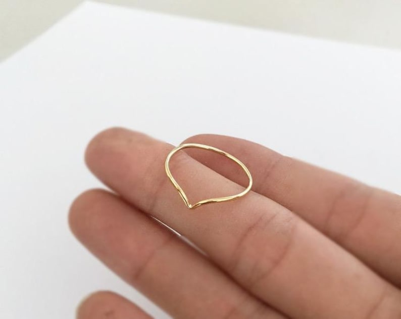 May include: A delicate gold-colored ring with a V-shaped design. The ring is thin and appears to be made of a single piece of metal. The ring is shown on a person's finger against a white background.