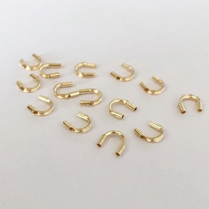 Op de afbeelding: Een groep van 14 gouden metalen sprongringen, ook wel bekend als jump rings, liggen verspreid op een wit oppervlak. De sprongringen zijn allemaal even groot en even gevormd, en ze zijn allemaal open.