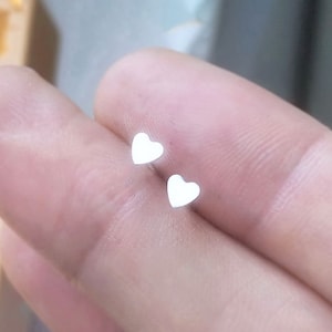 May include: A pair of small, white heart-shaped stud earrings. The earrings are shown on a person's finger.