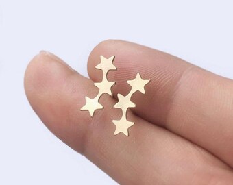 May include: Two gold-colored star-shaped earrings. Each earring features three stars connected in a vertical line. The earrings are small and delicate, suitable for everyday wear. The earrings are shown on a finger.
