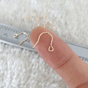 May include: Close-up of a gold-colored earring hook with a twisted design. The hook is held by a person's finger, with other hooks visible in the background. The background is a soft, white surface.