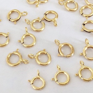 Könnte beinhalten: Eine Gruppe von 18 goldfarbenen Metall-Sprühringen. Die Sprühringe sind rund und haben einen federbelasteten Verschluss. Sie werden häufig in der Schmuckherstellung verwendet.