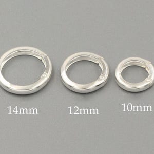 Könnte beinhalten: Drei silberfarbene runde Federringverschlüsse in verschiedenen Größen. Der größte ist 14 mm, der mittlere 12 mm und der kleinste 10 mm. Die Verschlüsse sind vor grauem Hintergrund abgebildet.