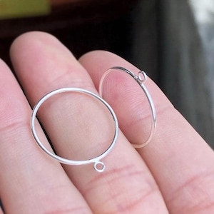 May include: Two silver-colored, circular jewelry findings with small loops for attaching to other components. The findings are held in a person's hand, and are designed for jewelry making. The findings are thin and delicate.