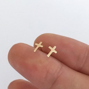 May include: A pair of small, gold-colored cross stud earrings. The earrings are displayed against a light background. The crosses have a simple, minimalist design, with a polished finish. The earrings are small and delicate.