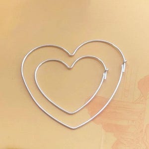 Pode incluir: Dois brincos de argola prateados em forma de coração. Um coração menor está aninhado dentro de um maior. Os brincos são feitos de metal fino e estão sobre um fundo castanho claro.