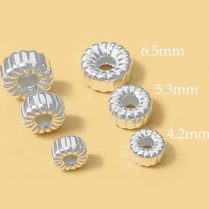 Könnte beinhalten: Silberfarbene, gerippte Perlen in verschiedenen Größen, darunter 6,5 mm, 5,3 mm und 4,2 mm. Die Perlen haben eine Donut-Form mit einem zentralen Loch. Der Hintergrund ist einfarbig hellgelb.