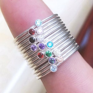 May include: A selection of silver rings, each featuring a different coloured gemstone. Gemstones include red, black, purple, green, and clear. The rings are stacked on a finger, displaying the variety of colours and designs.