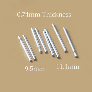 Könnte beinhalten: Auf beigem Hintergrund sind mehrere silberfarbene Metallstäbe unterschiedlicher Länge zu sehen. Der Text gibt eine Dicke von 0,74 mm an, mit Längen von 9,5 mm und 11,1 mm. Die Stäbe sind zylindrisch mit einem leicht breiteren Ende.