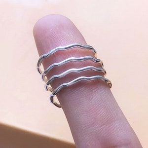 Może przedstawiać: Zestaw czterech srebrnych pierścionków w kształcie fali, ułożonych na palcu. Pierścionki są cienkie i mają falisty wzór.