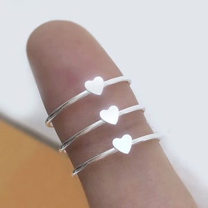 Puede incluir: Tres anillos de plata con un diseño en forma de corazón en cada anillo. Los anillos están apilados uno encima del otro en un dedo.