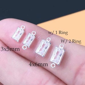 Puede incluir: Cuatro dijes rectangulares de tono plateado con piedras facetadas transparentes. Dos dijes miden 3x5mm con una argolla, y dos miden 4x6mm con dos argollas. El texto w/ 1 Ring, W/ 2 Ring, 3x5mm y 4x6mm está impreso cerca de los componentes.