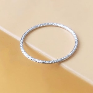 Puede incluir: Un anillo fino de plata con un diseño texturizado y retorcido. El anillo se presenta sobre un fondo naranja claro. El anillo parece ser de metal.