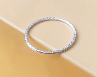 May include: A thin, silver ring with a textured, twisted design. The ring is set against a light orange background. The ring appears to be made of metal.