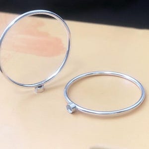 May include: Two silver-colored bangles. One bangle is upright, supported by a small, round base. The other bangle lies flat. Both have a simple, minimalist design, with a small, round accent on one side. The background is a light tan color.